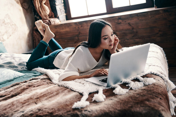 Relaxing at home. Beautiful young woman using laptop while lying on the bed at home - Fotoğraf, Görsel