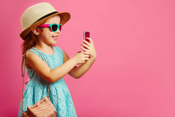 Beautiful little girl with charming smile looks at phone, has a good mood, dressed in fashionable blue dress, wearing a beach hat and sunglasses, standing on isolated pink. - Photo, Image