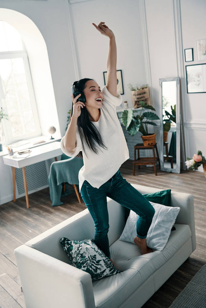 Superstar inside her. Full length of attractive young woman wearing headphones and smiling while dancing on sofa at home - Fotoğraf, Görsel