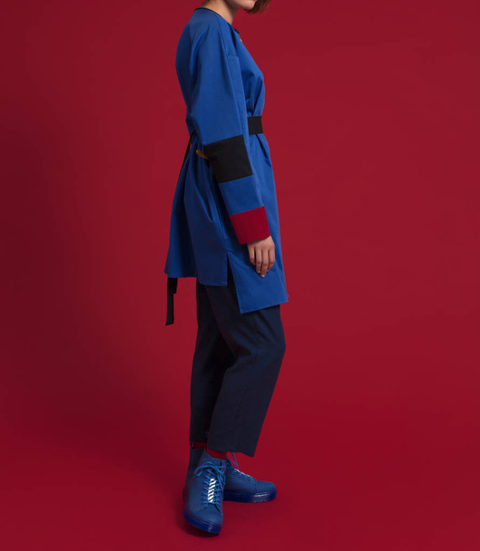 Ένα κορίτσι σε ένα όμορφο, μοντέρνο, πολυτελές μπλε πουκάμισο ρόμπα με λευκή τσέπη και πολύχρωμα μανίκια, μπλε παντελόνια, μπλε παπούτσια, κόκκινες κάλτσες σε κόκκινο φόντο - Φωτογραφία, εικόνα