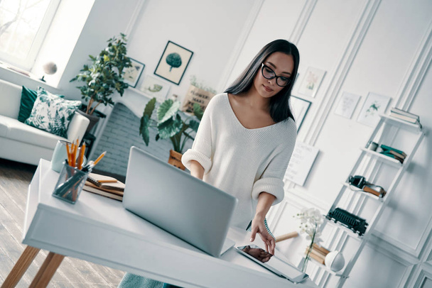 Kaunis nuori nainen käyttää kannettavaa tietokonetta työskennellessään kotitoimistossa
  - Valokuva, kuva