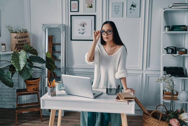 Smart and confident woman looking at camera and adjusting eyewear while standing in home office           - Foto, Bild