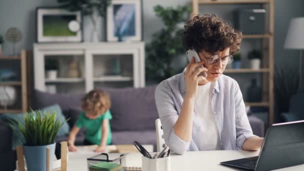 Angry freelancer talking on phone using laptop while her son playing in room - Video