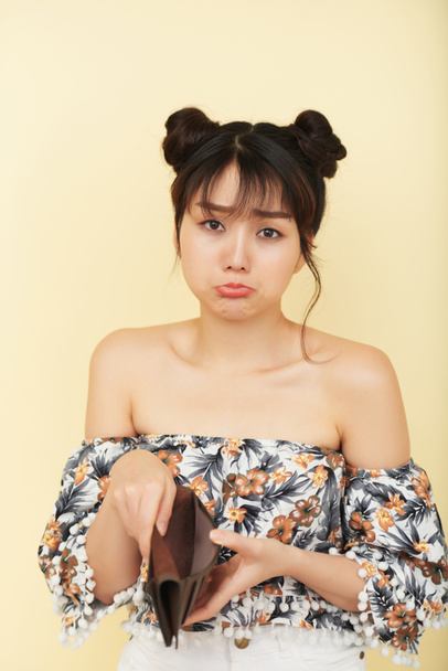 charmante junge stilvolle asiatische Frau blickt traurig in die Kamera und zeigt leere Brieftasche auf gelbem Hintergrund - Foto, Bild