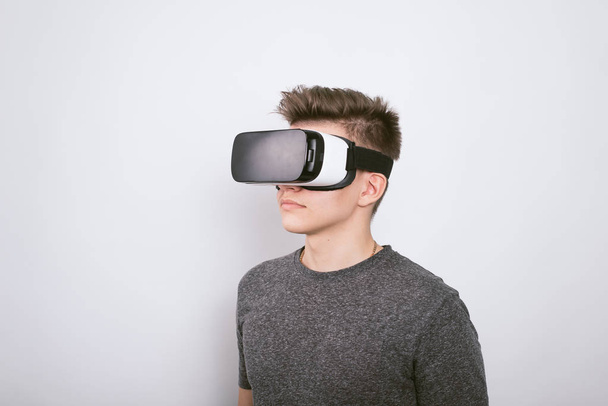 Un chico joven, un adolescente está de pie en un villard vr gafas sobre un fondo blanco. Realidad aumentada en nuestro tiempo. Un niño de gafas de realidad virtual juega y aprende en la escuela
. - Foto, imagen