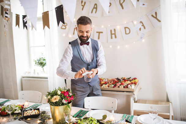 Mature man with bow and vest setting a table for an indoor party. - Foto, Bild