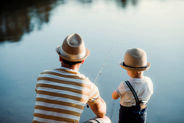 A rear view of mature father with a small toddler son outdoors fishing by a lake. - 写真・画像