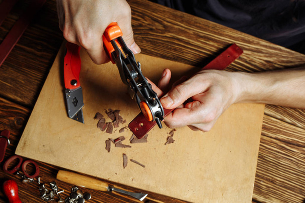 Maestro sosteniendo un agujero y un pedazo de cuero. Sobre mesa de madera marrón dispersa con herramientas y accesorios
. - Foto, Imagen