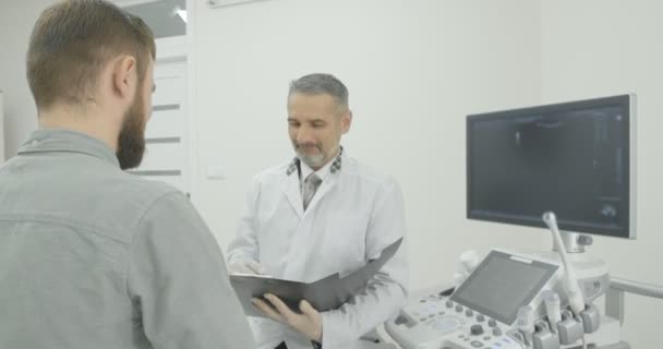 Mladý muž nosí v šedé košili po poradě se svým terapeutem. Pacient se dívá na doktora a poslouchá. Dospělý lékař, oblečený do bílé lékařské uniformy, hledí dolů. - Záběry, video