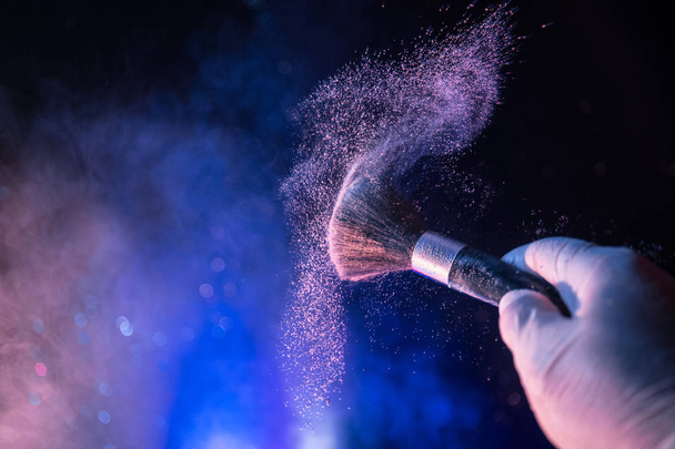 Makeup brush in hand with cosmetic powder on dark background with light and smoke. Powder splash on dark. Selective focus - Foto, imagen