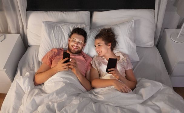 happy couple using smartphones in bed at night - Foto, afbeelding