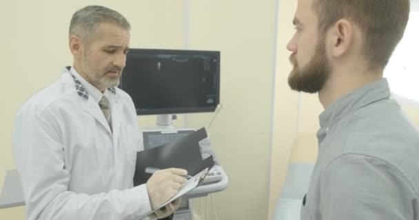 Jeune homme vêtu d'une chemise grise en consultation avec son thérapeute. Patient regardant le docteur, écoutant. Médecin mature portant en uniforme médical blanc regardant vers le bas
. - Séquence, vidéo