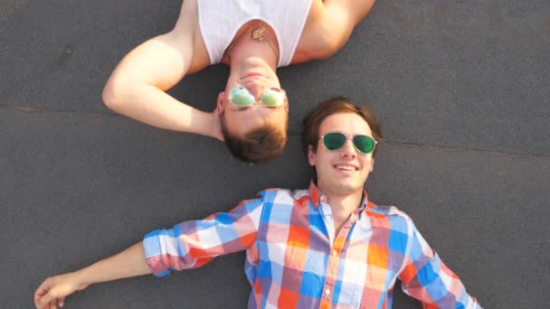 Bovenaanzicht van twee knappe homo's in zonnebril liegen met geluk en vreugde uitdrukking op het gezicht. Gelukkig jongemannen glimlachend en genieten van het leven samen. Vrienden buiten ontspannen. Slow motion close-up - Video