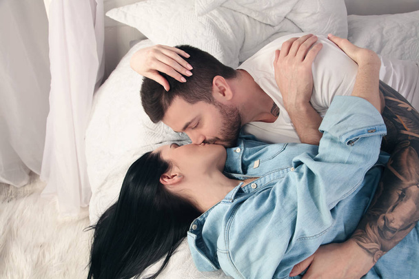 Χαρούμενο ζευγάρι 30s αγκάλιασμα και το φίλημα μαζί, ξαπλωμένοι στο κρεβάτι στο σπίτι ή το ξενοδοχείο διαμέρισμα - Φωτογραφία, εικόνα