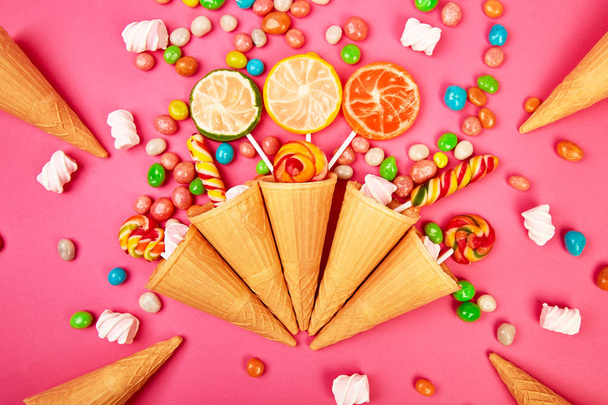Ice cream waffles cones with colorful candy, sweets jelly, lollipop on stick, scattering of multicolored sweets on pink background. Flat lay, Top view. Copy space. - 写真・画像