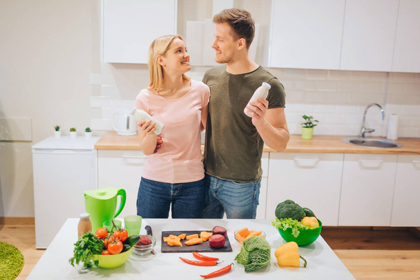 Vegan happy loving couple holds bottles with natural smoothie while cooking raw vegetables in white kitchen. Diet detox. Happy family. Raw food diet. Vegetarian food. - Φωτογραφία, εικόνα