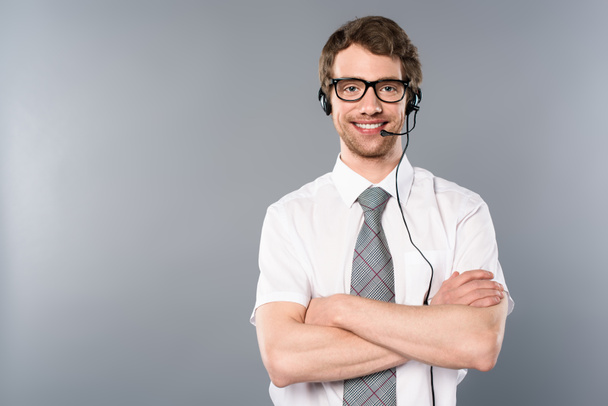 Lächelnder Callcenter-Betreiber mit Brille und Headset mit verschränkten Armen auf grauem Hintergrund - Foto, Bild