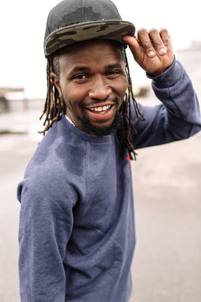 Handsome African American with dreadlocks in brown trousers and a blue sweater in a black cap on his head against the background of houses, city - Foto, afbeelding
