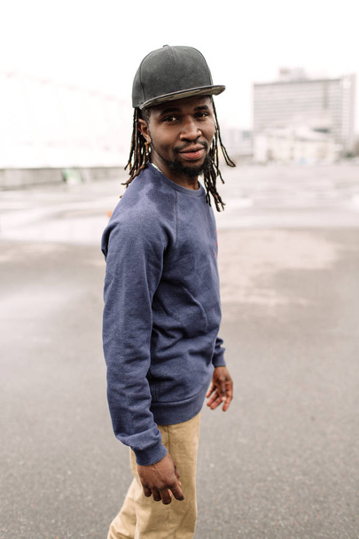 Handsome African American with dreadlocks in brown trousers and a blue sweater in a black cap on his head against the background of houses, city - Zdjęcie, obraz
