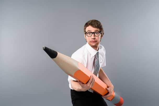 tense businessman in glasses holding huge and heavy decorative pencil on grey background - Zdjęcie, obraz