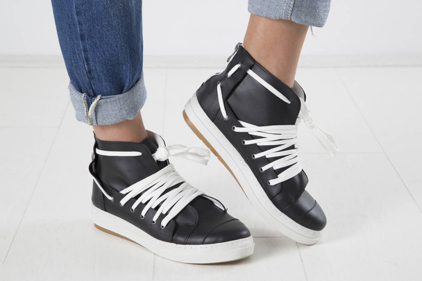 Studio Shot Of Female Legs In Stylish Black Leather Sneakers With White Shoelace, White Background Close Up - Foto, imagen