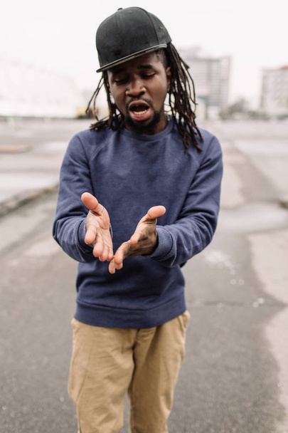 Handsome African American with dreadlocks in brown trousers and a blue sweater in a black cap on his head against the background of houses, city - Foto, Bild