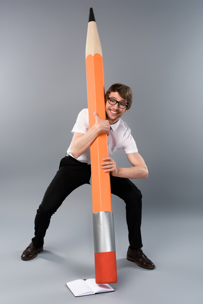 Улыбающийся бизнесмен в очках держит огромный декоративный карандаш рядом с блокнотом на сером фоне
 - Фото, изображение
