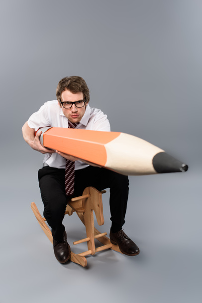 funny focused business man in glasses holding huge decorative pencil while riding rocking horse on grey background - Photo, Image