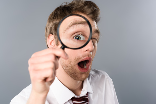 shocked businessman with funny face expression looking in magnifier on grey background - Zdjęcie, obraz