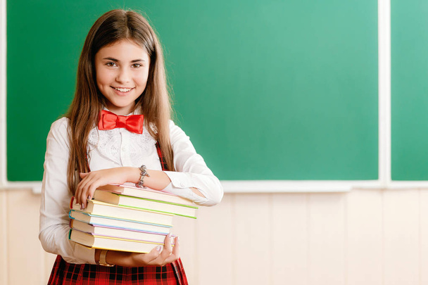 Μαθητή σε σχολική στολή, στέκεται σε μια σχολική τάξη απέναντι από το μαυροπίνακα και να κρατά τα σχολικά βιβλία - Φωτογραφία, εικόνα