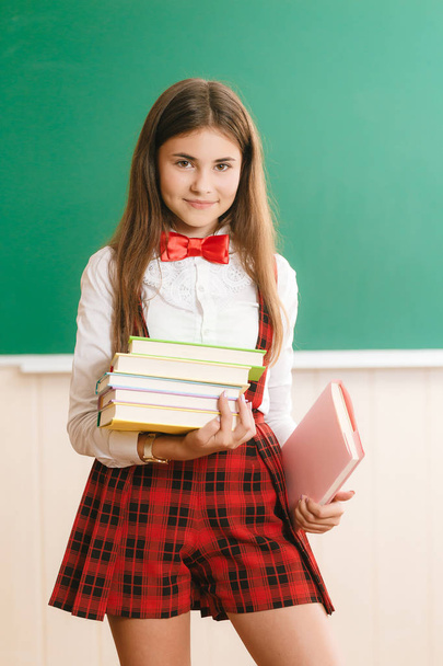 Alumno con uniforme escolar de pie en una clase escolar frente a la pizarra y sosteniendo libros de texto
 - Foto, imagen