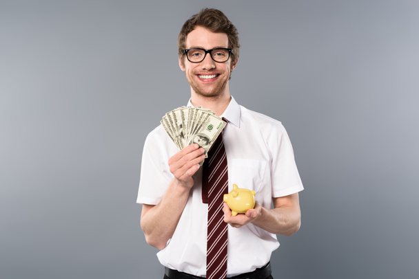 smiling businessman in glasses holding piggy bank and dollar banknotes on grey background - Foto, afbeelding