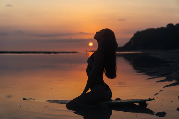 Silueta chica sexy en traje de baño acostado y posando en la tabla de surf en la playa durante la puesta del sol
 - Foto, Imagen