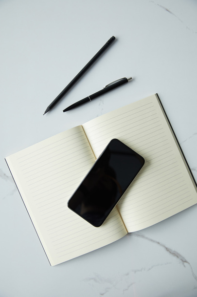 top view of smartphone with blank screen, notebook, pen and pencil on white marble surface - Photo, Image