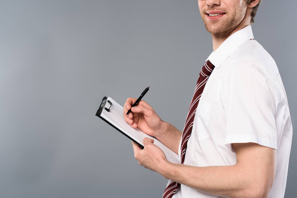 cropped view of smiling businessman man with pen writing on clipboard on grey background - Foto, afbeelding
