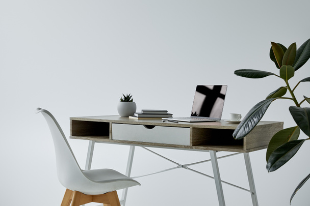 table with laptop, books, flowerpot, and white chair on grey background - Fotó, kép