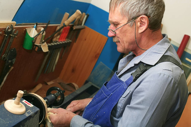 Чоловік пенсійного віку в окулярах і вуса обробляє дерев'яну заготовку на токарному верстаті. Робоче місце тесляра
 - Фото, зображення