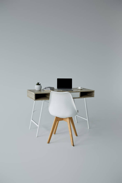 valkoinen tuoli, pöytä kannettavalla tietokoneella, kirjat ja kukkaruukku harmaalla pohjalla
 - Valokuva, kuva