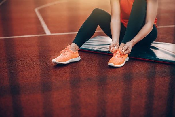 Immagine ritagliata di donna caucasica in abbigliamento sportivo seduta sul tappeto sul campo e allacciatura lacci delle scarpe. Ogni giorno è un buon giorno per l'allenamento
. - Foto, immagini