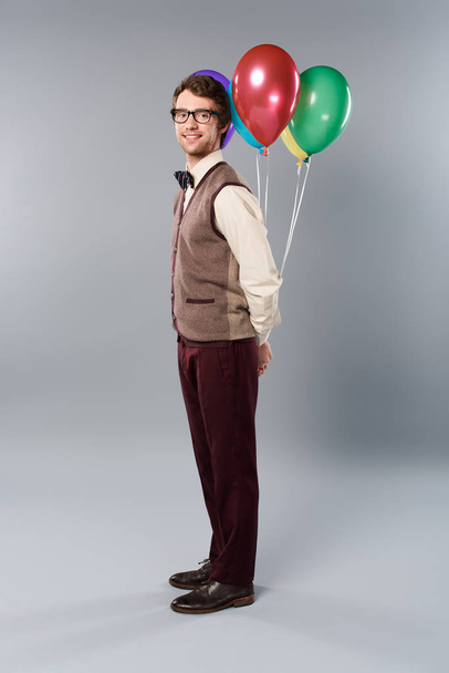 positive man in glasses holding multicolored balloons on grey background - Fotoğraf, Görsel