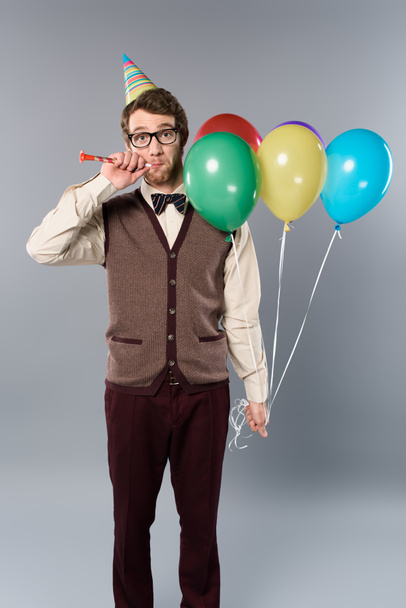 funny man in glasses and party cap holding multicolored balloons and party horn on grey background - Photo, image