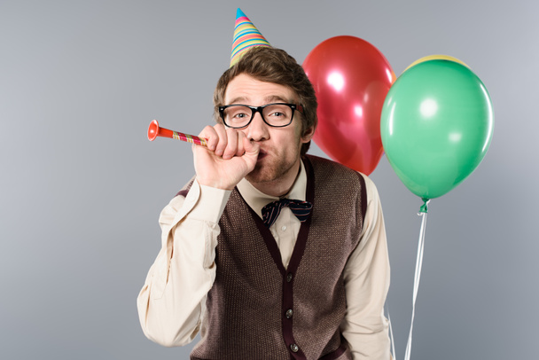 funny man in glasses and party cap holding multicolored balloons and blowing in party horn on grey background - Foto, afbeelding