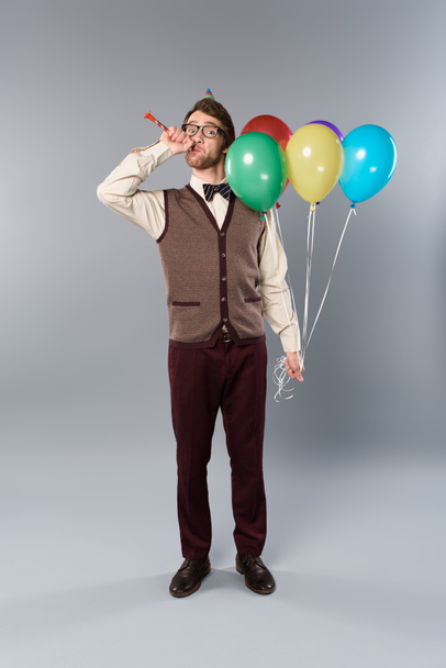 man in glasses and party cap holding multicolored balloons and blowing in party horn on grey background - Fotoğraf, Görsel