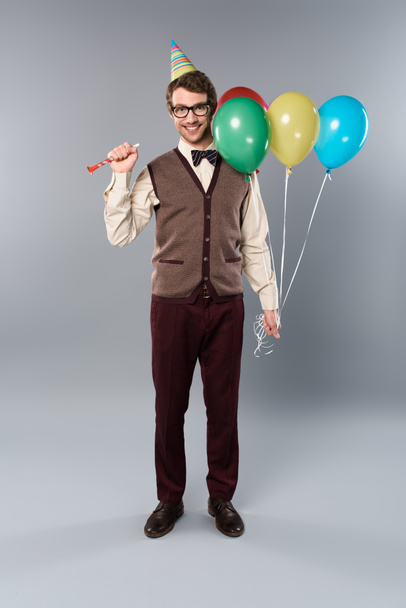 happy man in glasses and party cap holding multicolored balloons and party horn on grey background - Foto, Imagem