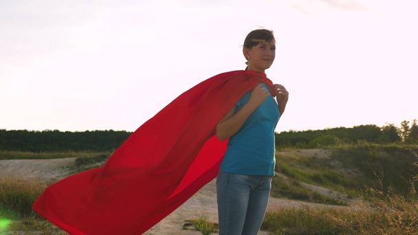 bela menina super-herói em pé no campo em um manto vermelho, vacilante manto no vento. Movimento lento. Uma jovem sonha em se tornar uma super-heroína. menina caminha em um manto vermelho expressão de sonhos
 - Foto, Imagem