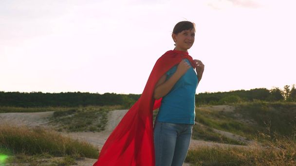 beautiful girl superhero standing on the field in a red cloak, cloak fluttering in the wind. Slow motion. A young girl dreams of becoming a superhero. girl walks in a red cloak expression of dreams - Photo, Image