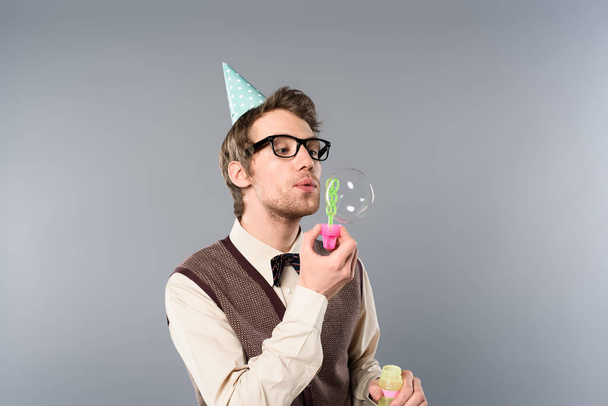 man in vintage clothing and party cap blowing soap bubbles on grey background - Foto, Imagem