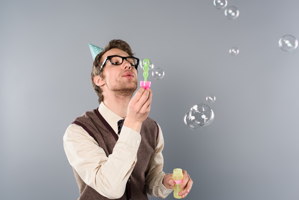 adult man in vintage clothing and party cap blowing soap bubbles on grey background - Photo, Image