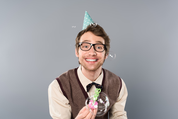 smiling man in vintage clothing and party cap blowing soap bubbles on grey background - Photo, Image