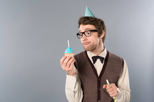 man in glazen en partij cap met grappige gezicht expressie holding cupcake met kaars  - Foto, afbeelding